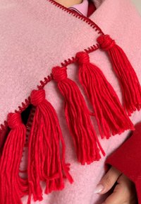 Tissu rose doux avec des pompons en fil rouge décoratif le long du bord, tenu par une main portant une manche rouge.