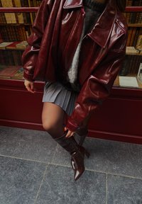 Bordo odos oversize švarkas, pilka klostuota sijonas ir rudi aukštakulniai batai su smailiu nosimi ant tekstūruoto pilko grindų paviršiaus.