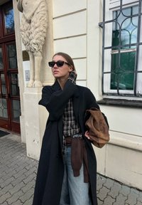 Femme en manteau noir et lunettes de soleil ajuste ses cheveux, tenant un sac marron, debout près d'un bâtiment beige avec des jambes de lion sculptées et une fenêtre à barreaux.