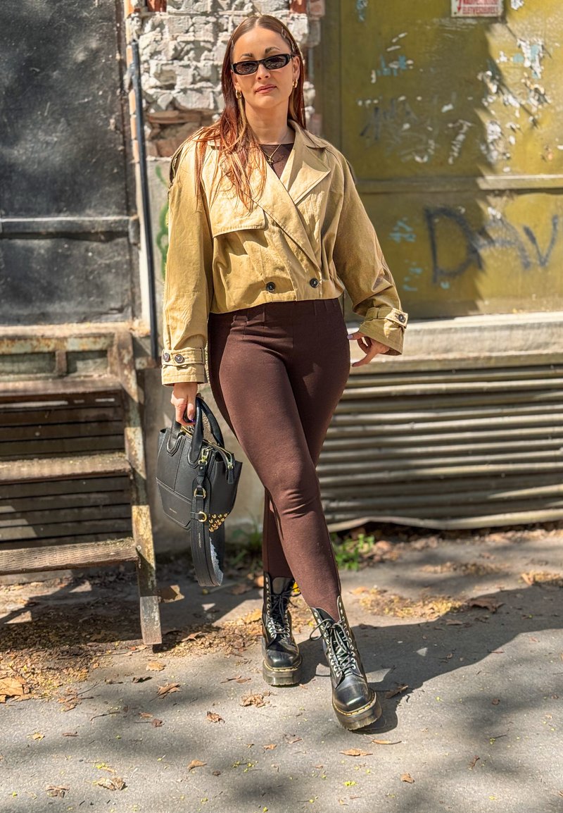 Femme aux longs cheveux portant des lunettes de soleil, une veste courte beige, des leggings marron, des bottes noires, tenant un sac à main noir, debout à l'extérieur près des escaliers.