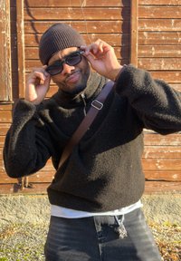 Homme portant un bonnet brun et un pull noir ajuste des lunettes de soleil noires devant un mur en bois, avec une sangle traversant la poitrine et des clés suspendues au jean.