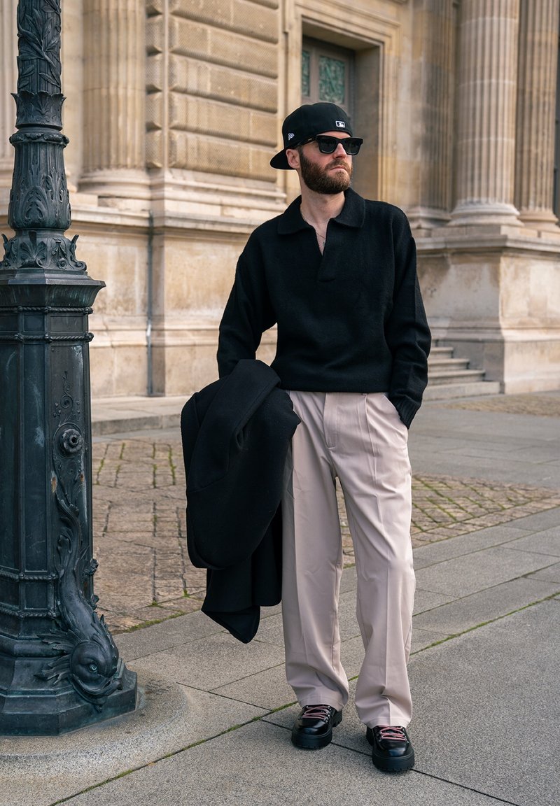 Homme barbu portant une chemise noire, un pantalon beige, des baskets et une casquette à l'envers, tenant un manteau noir, debout dans une rue de la ville près d'un lampadaire orné.