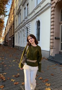 Zielony oversize'owy sweter, biała warstwowa bluzka, kremowe spodnie z szerokimi nogawkami oraz wzorzysty pasek na biodra. W tle liście i brukowane kamienie.