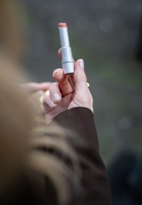 MAC leppestift i en sølvmetallisk tube, med en myk nude nyanse synlig. Holdt i en hånd med nøytral neglelakken, mot en uskarp bakgrunn.
