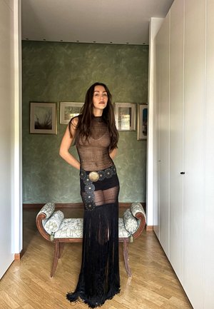 Žena s dlhými vlasmi oblečená v čiernych sieťových šatách a so širokým dekoratívnym opaskom, stojaca v miestnosti s drevenou podlahou a rámovanými umeleckými dielami.