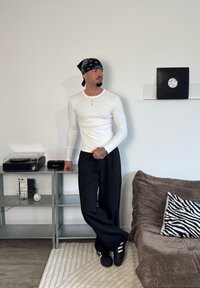 Biała koszula z długim rękawem, czarne spodnie i czarne trampki. Dodatki to bandana i warstwowe naszyjniki. W pokoju znajduje się gramofon.