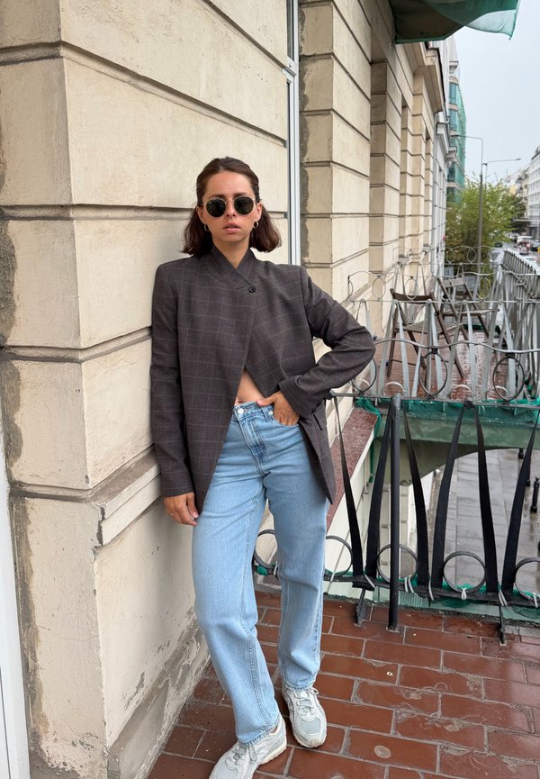 Femme portant un blazer sombre, un jean bleu clair, des baskets blanches et des lunettes de soleil rondes, debout sur un balcon à côté d'un mur beige.