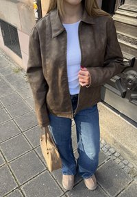 Brūna mākslīgās ādas jakas ar rāvējslēdzēju, apvienota ar baltu krekla un ziliem izplestiem džinsiem. Gaiši brūna rokassoma un gaiša apavi.
