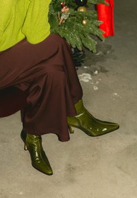 Personne portant un pull en maille vert, une jupe marron et des bottines vertes brillantes, assise près d'un sapin de Noël décoré sur un sol en béton.
