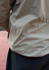 Jachetă ușoară bej cu textură netedă, având un logo pe partea stângă inferioară și o cusătură laterală. Purtată peste pantaloni închiși la culoare.