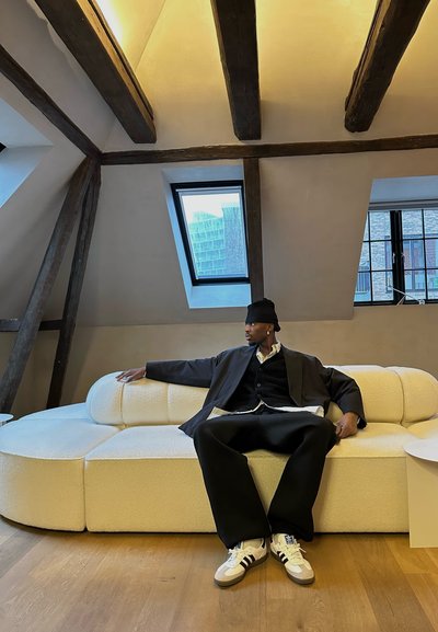 Sofá curvado tapizado en blanco con tela texturizada, ubicado sobre un suelo de madera; modelo vistiendo un abrigo negro y pantalones holgados, mostrando una postura relajada.