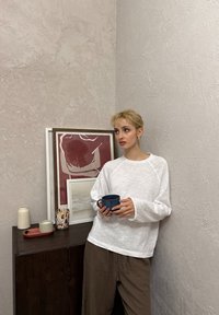 Giovane con capelli biondi corti, indossa una maglietta bianca e pantaloni marroni, tiene in mano una tazza blu e si trova vicino a un mobile in legno scuro con opere d'arte incorniciate e oggetti decorativi.