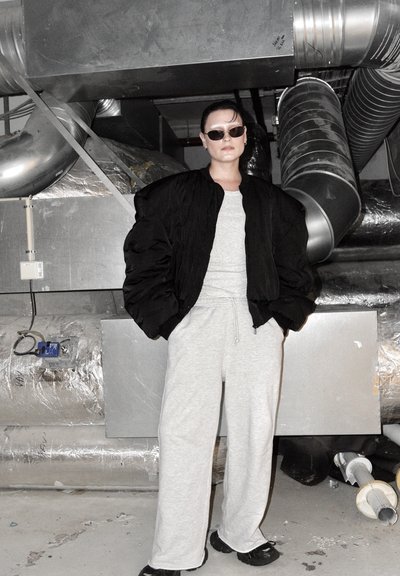 Chaqueta bomber negra acolchada, pantalones deportivos gris claro y zapatos oscuros. Fondo industrial con conductos y tuberías de metal expuestos.