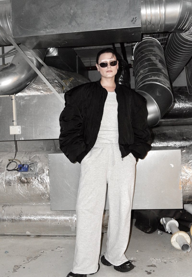 Doudoune noire, pantalon de survêtement gris clair, et chaussures foncées. Arrière-plan industriel avec conduits métalliques et tuyaux apparents.