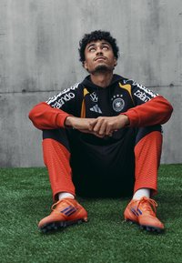 Μαύρη και κόκκινη φόρμα με λογότυπα και λεπτομέρειες; εφαρμοστό σχέδιο, υφασμάτινα μανίκια και παντελόνια με σχέδιο. Ποδόσφαιρικες παπούτσια πορτοκαλί με μπλε ρίγες.