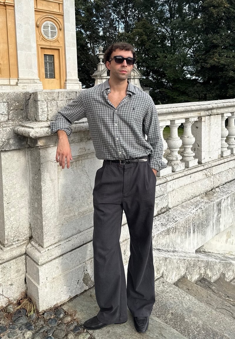 Uomo con occhiali da sole, camicia a scacchi grigia e pantaloni scuri, si appoggia a un parapetto di pietra vicino a un edificio storico, con alberi sullo sfondo.