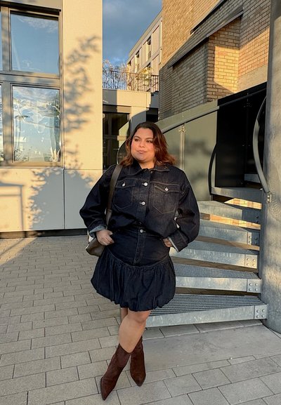 Mujer con chaqueta de mezclilla oscura y falda, botas marrones, de pie en un área pavimentada junto a un edificio moderno y una escalera de metal en espiral, bajo la luz del sol.