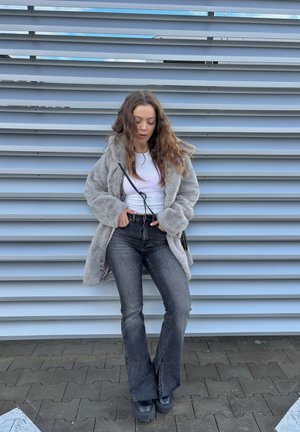 Abrigo de piel sintética gris, blusa blanca, vaqueros negros de tiro alto y pata de elefante, y botas negras de plataforma, de pie frente a un fondo de panel texturizado gris.