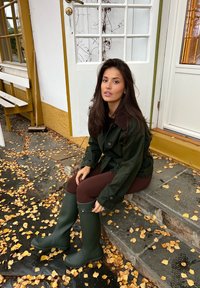 Donna con giacca verde e stivali di gomma verdi seduta su gradini di pietra fuori da una casa gialla e bianca con foglie autunnali cadute intorno.