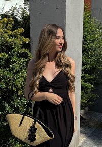 Jeune femme aux longs cheveux ondulés, vêtue d'une robe noire, tenant un sac cabas en paille, debout en plein air près de buissons verts et d'un mur gris clair.