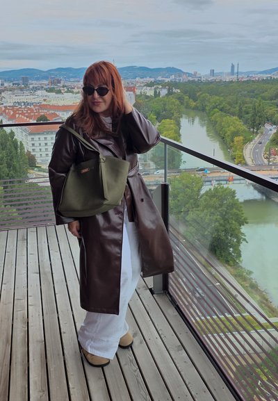 Abrigo largo de cuero marrón, pantalones blancos, calzado marrón y bolsa verde oliva. Gafas de sol negras y fondo urbano con vegetación y un río.