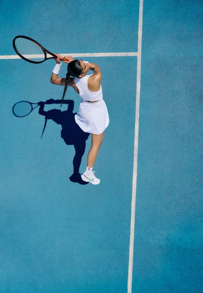 Jugadora de tenis con un vestido blanco en una cancha azul, sosteniendo una raqueta y proyectando una sombra. Lleva zapatillas blancas y calcetas hasta el tobillo.