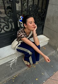 Haut à imprimé léopard, pantalon de jogging bleu marine avec des rayures dorées, chaussures marron, et un sac tressé avec des franges ; accessoire floral dans les cheveux.