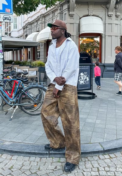 Pantalones sueltos de estampado marrón con cintura alta; camisa blanca de botones con rayas; gorra marrón; gafas de sol oscuras; zapatos negros. Entorno urbano al aire libre.