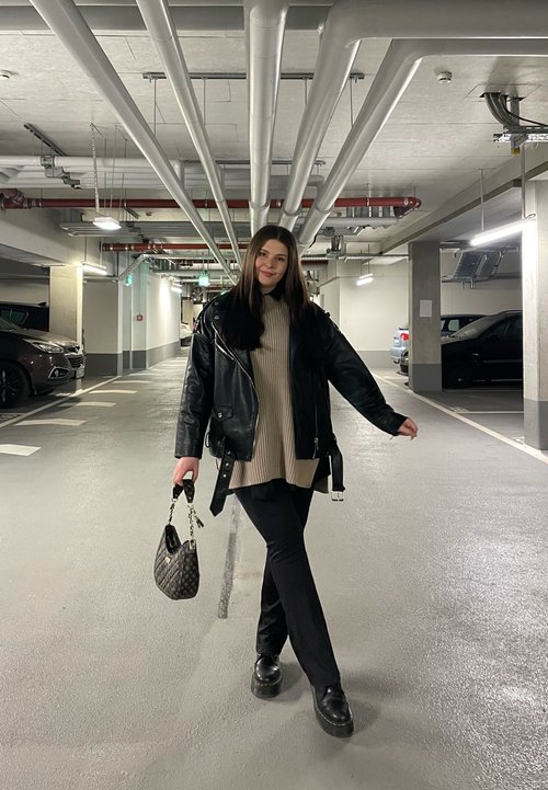 Crna kožna jakna preko bež rebraste majice, crne hlače i platforme, drži torbu sa preklopom, u garaži s cijevima iznad.