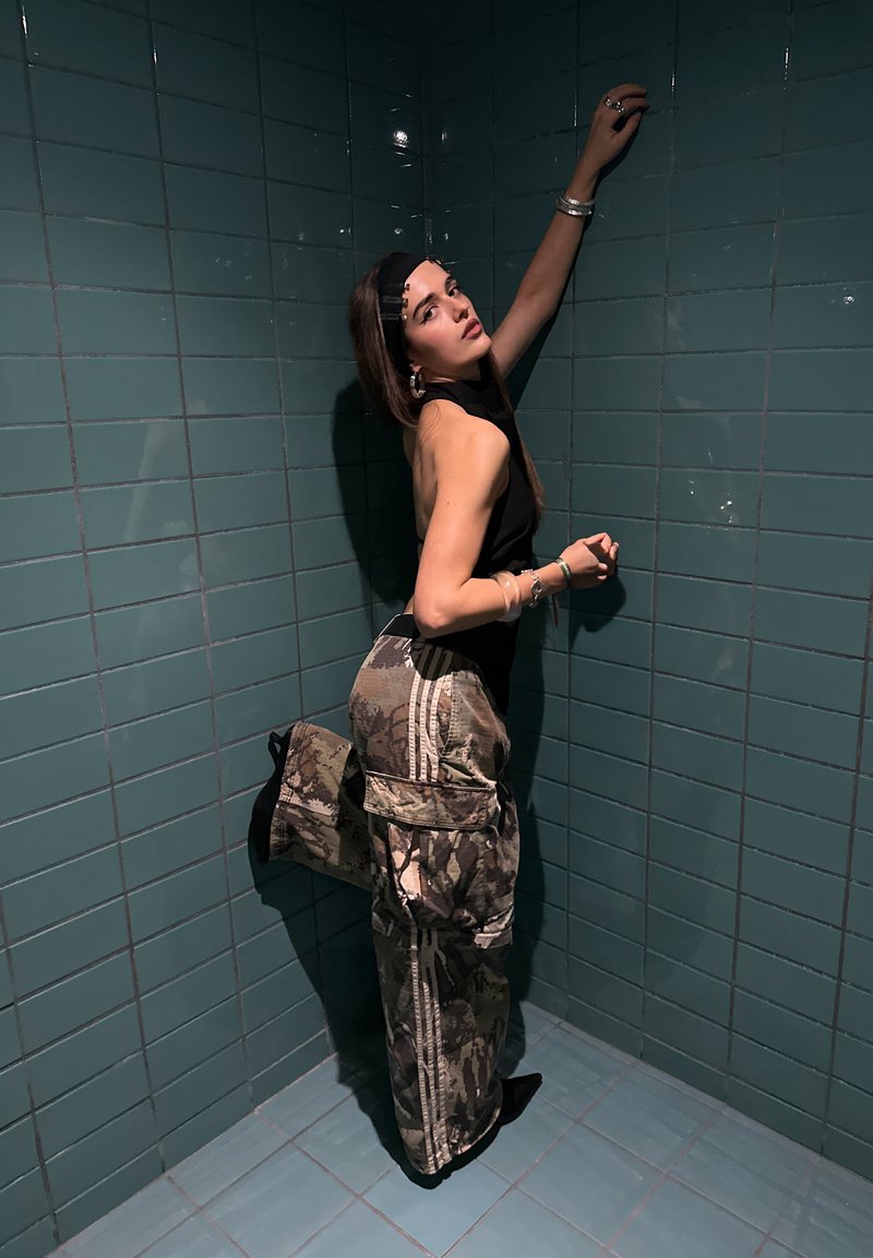 Top nero senza maniche con collo alto, pantaloni cargo mimetici con tasche e stivali neri lucidi con tacco. Sullo sfondo si vedono pareti piastrellate di turchese.