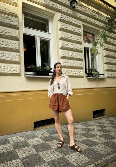 Camisa de rayas beige ligera, camiseta sin mangas blanca, pantalones cortos de lino color óxido y sandalias marrones tipo slide. De pie sobre adoquines cerca de una pared amarilla.