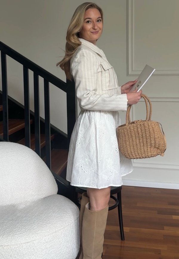 Femme en robe blanche et veste tenant un livre et un sac tissé, debout près d'une rampe d'escalier noire et d'une chaise blanche rembourrée.