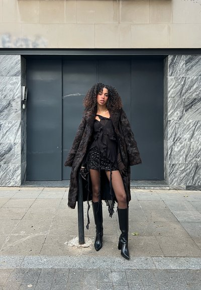 Un abrigo negro de piel sintética sobre un top negro con volantes y una minifalda con lentejuelas, combinado con botas negras hasta la rodilla; entorno urbano con un fondo de mármol.