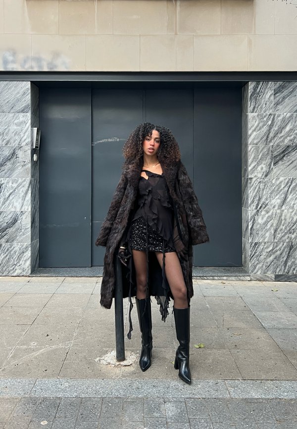 En svart fuskpälsjacka över en svart topp med volanger och en paljettprydd minikjol, kombinerat med knähöga svarta stövlar; stadsmiljö med en marmorbakgrund.