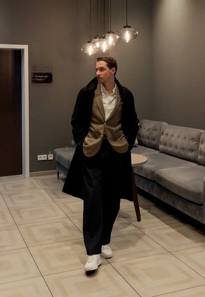 Hombre vestido con un abrigo negro sobre un blazer marrón, camisa gris y pantalones de rayas. Zapatillas blancas; interior con sofá gris y luces colgantes.