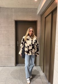 Bézs és fekete műszőrme dzseki boxy szabással, világoskék farmerrel és fehér tornacipővel, egy lift mellett állva.