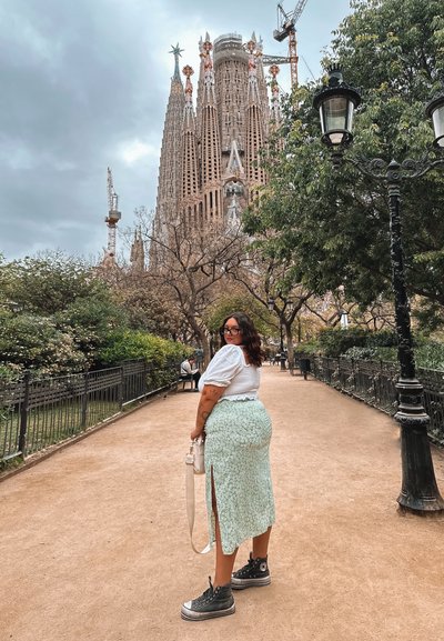 Mujer vestida con una blusa blanca y una falda floral verde menta con una gran abertura, zapatillas negras, de pie en un parque con la Sagrada Familia al fondo.