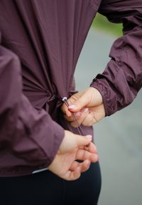Veste légère bordeaux avec un ourlet à cordon de serrage, présentant des poignets élastiques. Des mains ajustent le cordon à l'arrière pour un meilleur ajustement.