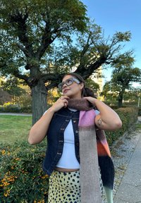 Flerfarget ullskjerf med rosa, oransje og grå seksjoner drapert rundt halsen. Har en denimvest og mønstret skjørt.