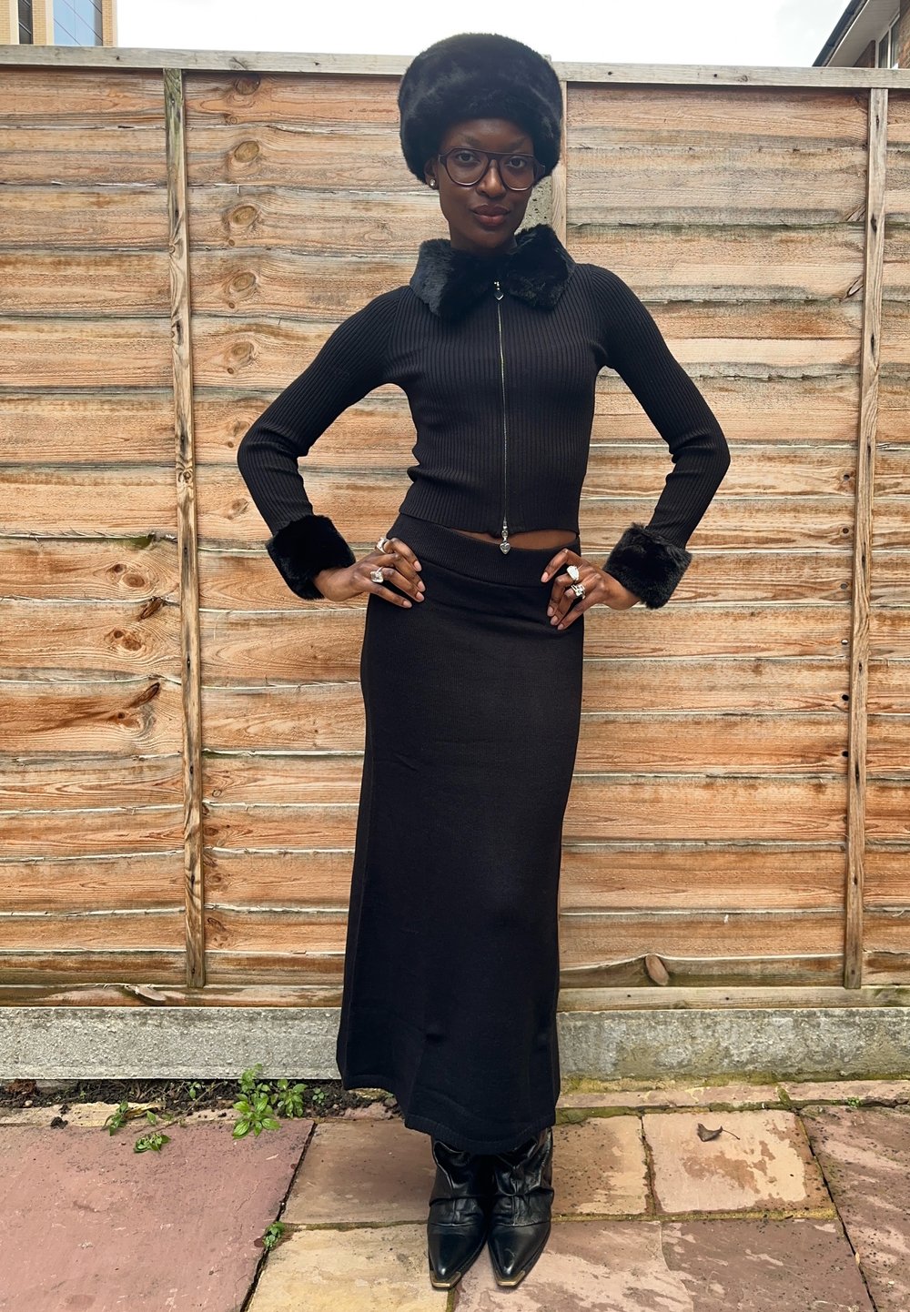Čierny rebrovaný dvojdielny outfit pozostávajúci z priliehavého topu na zips s umelým kožúškom na golieri a manžetách a zodpovedajúcej dlhej sukne. Čierne špicaté čižmy.