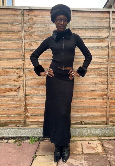 Conjunto negro de dos piezas con textura acanalada, que incluye un top ajustado con cremallera y cuello y puños de piel sintética, y una falda larga a juego. Botas negras puntiagudas.