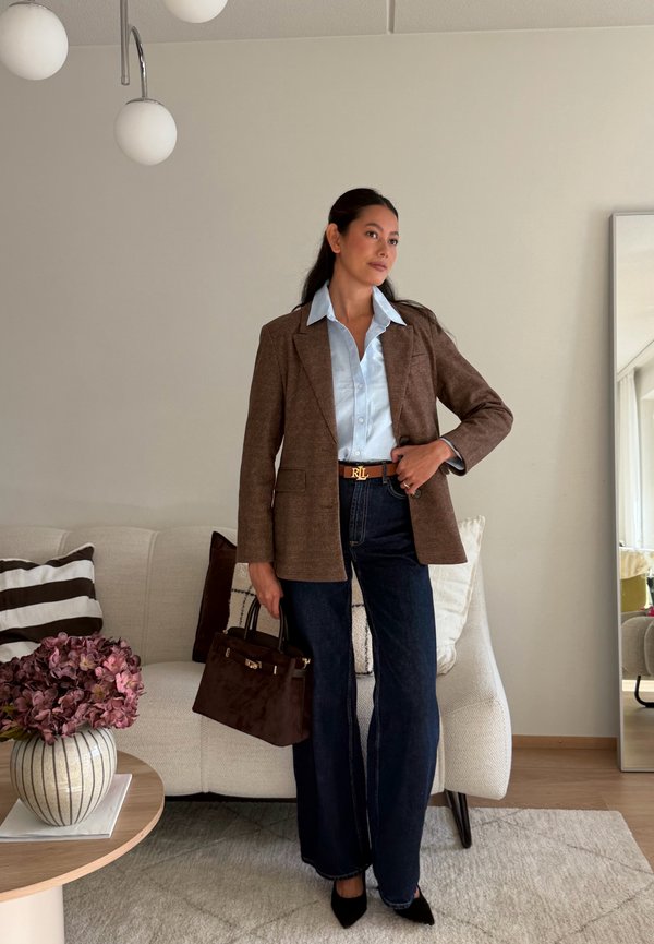 Una mujer lleva un blazer marrón, una camisa azul pálido, vaqueros oscuros y zapatos negros, sosteniendo un bolso marrón en un salón.