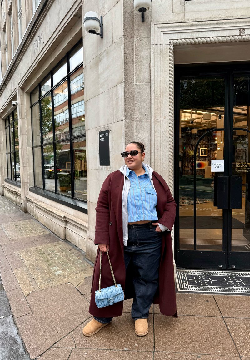Un long pardessus bordeaux, une chemise bleu clair rayée, un sweat à capuche gris, un jean foncé, des bottes couleur fauve et un sac à main matelassé bleu. Environnement urbain avec des portes vitrées.