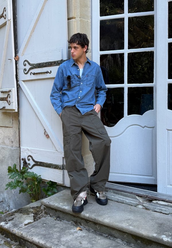 Joven de pie sobre escalones de piedra delante de una puerta blanca con cristales, viste camisa vaquera azul, pantalones cargo color oliva y zapatillas negras y blancas.