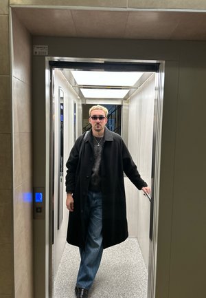 Interior de un ascensor con paredes grises y techo iluminado. Una persona con un abrigo negro y gafas de sol está en la entrada, mirando hacia adelante.