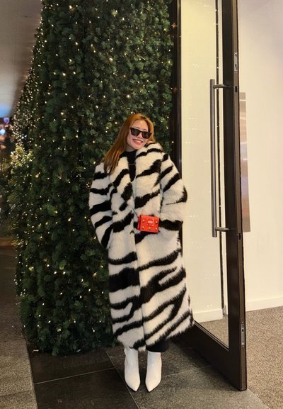 Mujer con un abrigo de piel a rayas blancas y negras y botas blancas, sonriente junto a un árbol de Navidad alto iluminado y una puerta de cristal abierta.