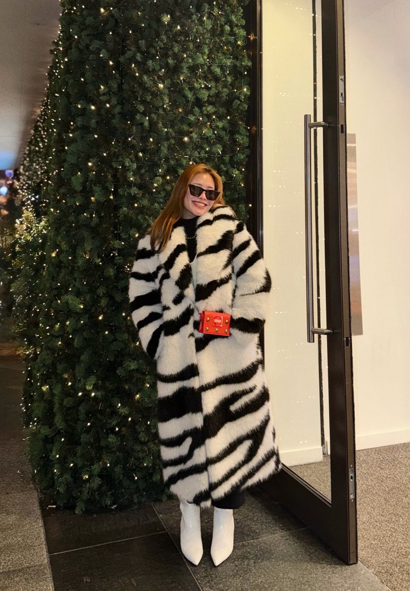 Eine Frau in einem schwarz-weiß gestreiften Pelzmantel und weißen Stiefeln steht lächelnd neben einem hohen, beleuchteten Weihnachtsbaum und einer offenen Glastür.