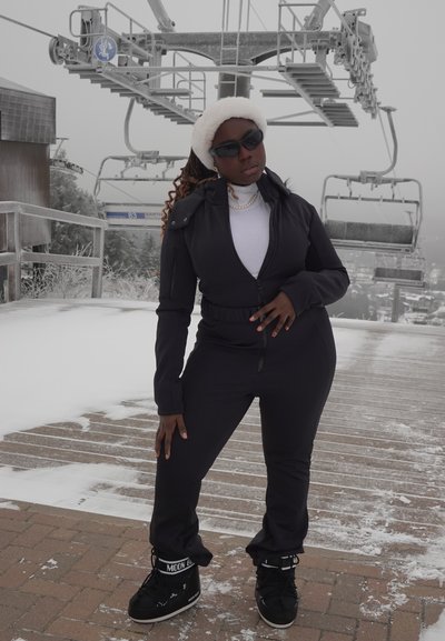 Chándal negro con cremallera y cintura ajustada, debajo un cuello alto blanco, y diadema de piel sintética blanca. Botas de nieve negras con detalles estampados.