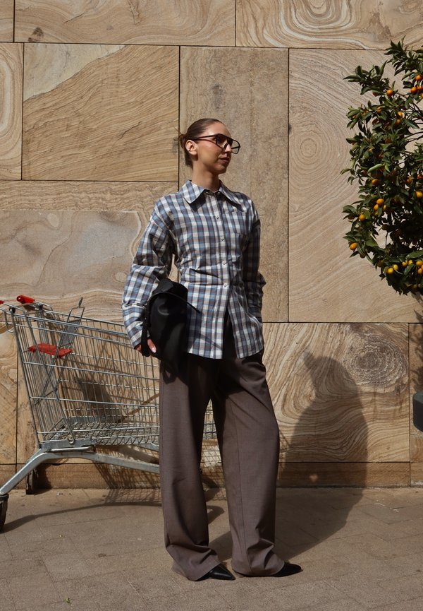 Personne avec des lunettes portant une chemise à carreaux et un pantalon large se tient à côté d'un chariot de supermarché et d'un oranger contre un mur en pierre.