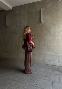 Γυναίκα με μακριά ξανθά μαλλιά, φορώντας γυαλιά ηλίου, μπορντό σακάκι και καφέ παντελόνι, που στέκεται μπροστά σε μια απλή τσιμεντένια τοιχογραφία με μια μεγάλη τσάντα.